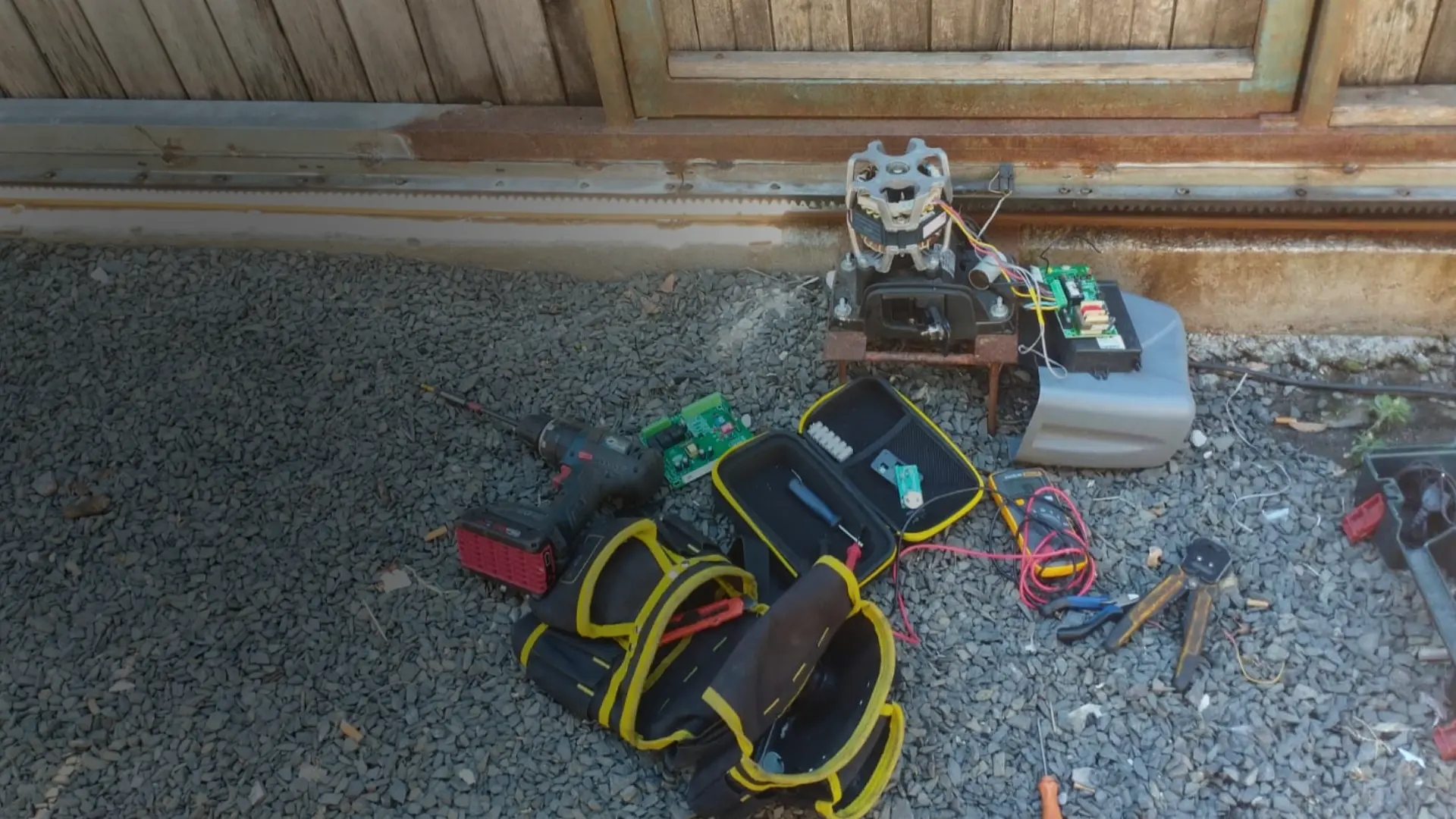 Motor de portão eletrônico desmontado no chão com ferramentas técnicas, multímetro e placa de comando, durante serviço de conserto.