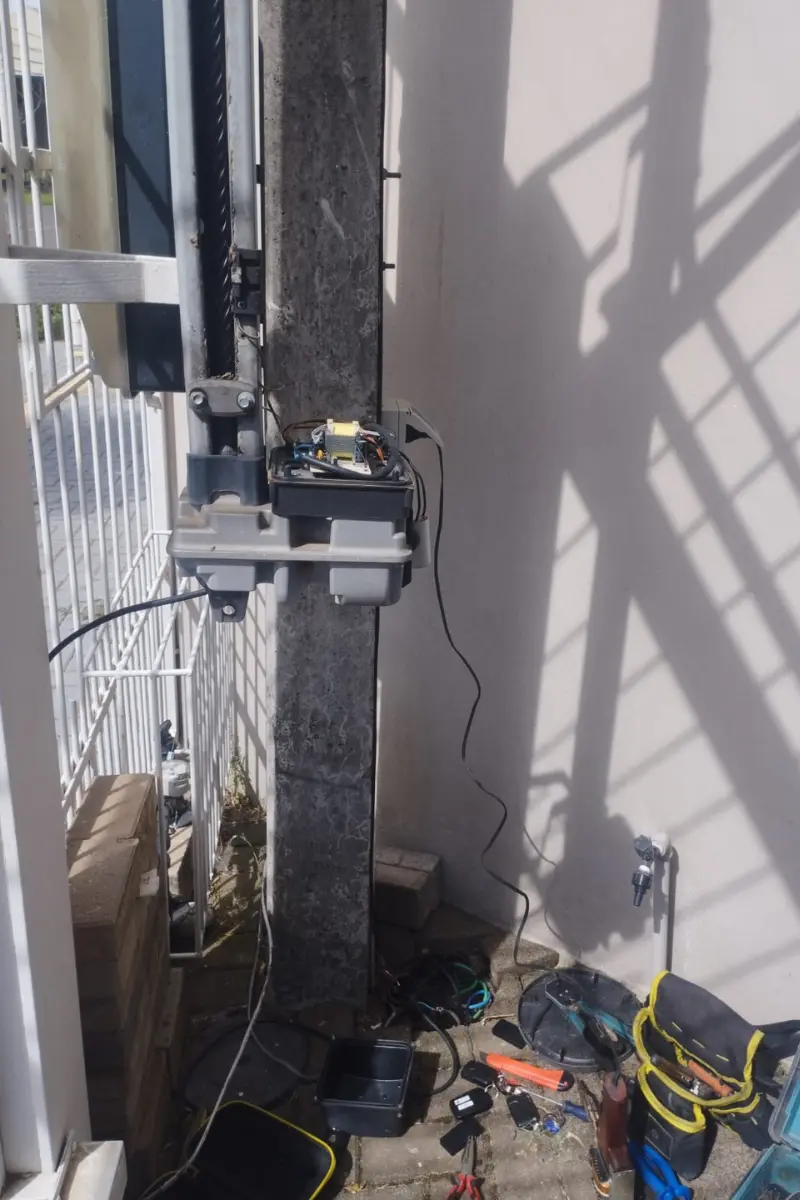 Motor de portão eletrônico basculante fixado em um pilar de concreto, com fiação aparente e ferramentas de manutenção técnica distribuídas no chão.