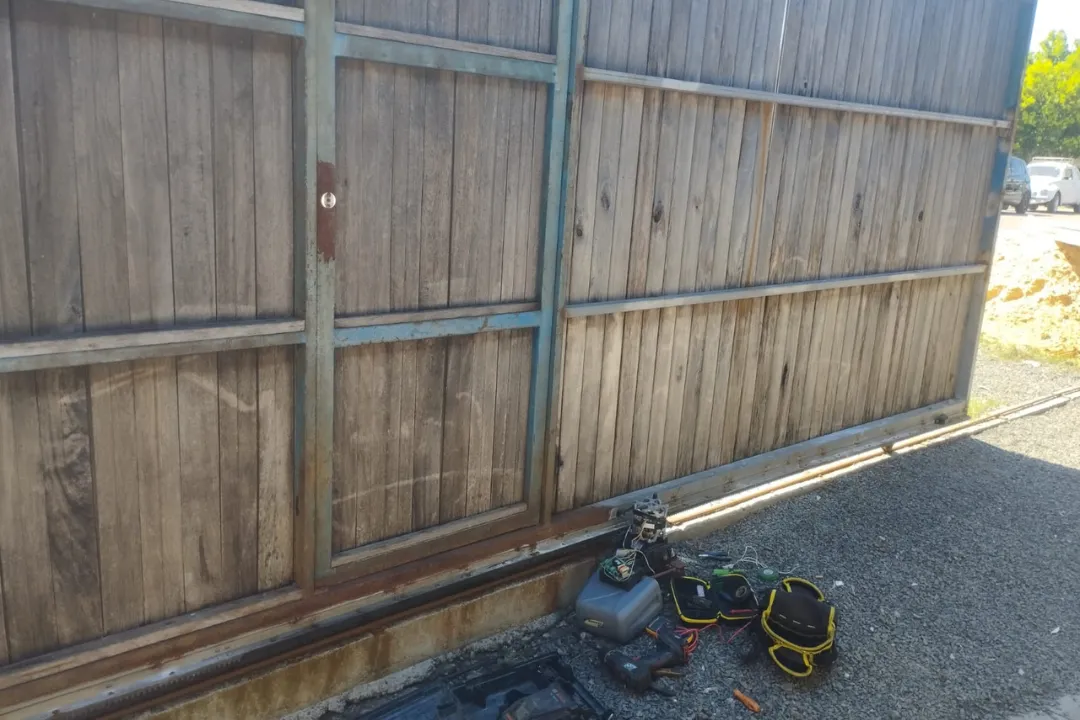 Ferramentas de técnico e peças de motor de portão eletrônico no chão durante conserto de portão deslizante de madeira.